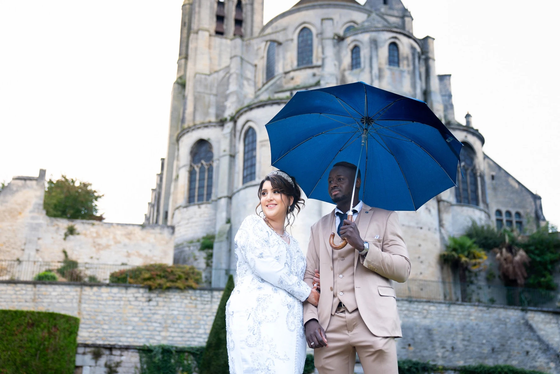 Un couple de mariés sous un parapluie bleu devant une église — Ziris Studio
